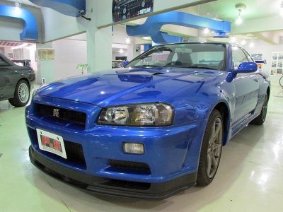 BNR34GT-R〔車台番号010107〕<br>
先代GT-Rでの反省点と、その開発過程で得られた技術的なノウハウを投入し「世界一の走り」をテーマに掲げて開発された10代目のR34型は、居住性よりも走行性能を重視し平成10年5月に登場。ボディーのサイズダウンを図ると共に剛性を大幅にアップ。走行安定性を向上させ、その重さを意識させない仕上りとなった。そして翌年1月にはGT-Rが発売され、Vスペック・VスペックⅡ・Mスペック...と開発陣の限りないエネルギーと情熱を注ぎ込まれ、常に「究極」を追い求め進化し続けてきた。そうGT-Rは決してとどまることを許されなかった。それらGT-Rの集大成ともいえる最終限定車VスペックⅡ「Nur」は発表当日に1000台が完売する快挙を成し遂げ、改めてスカイラインの人気の高さを証明しながら二世代GT-Rはフィナーレを迎えた。R34は20世紀最後のGT-Rとなったのである。この車両は、日産村山工場で最後に生産された、まさに記念すべき一台である。